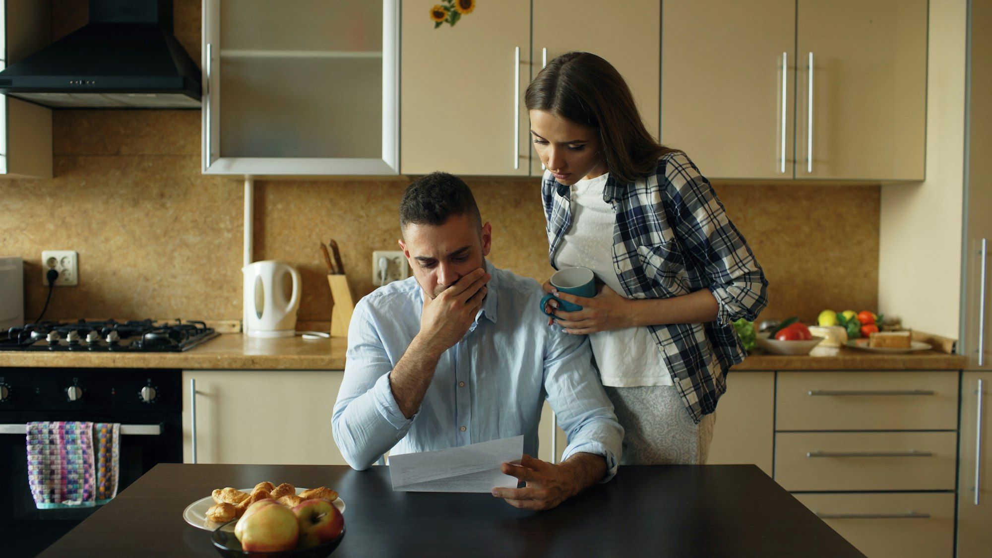 Worried man reading overdue bills at the kitchen table while his partner puts an arm around him