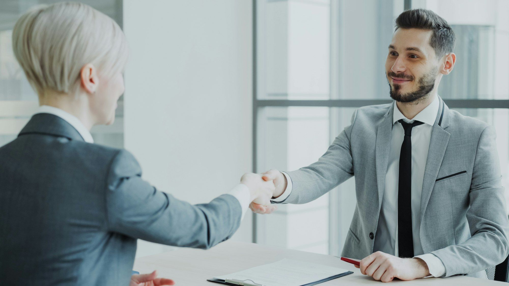 Two people shaking hands across a table in a bright office